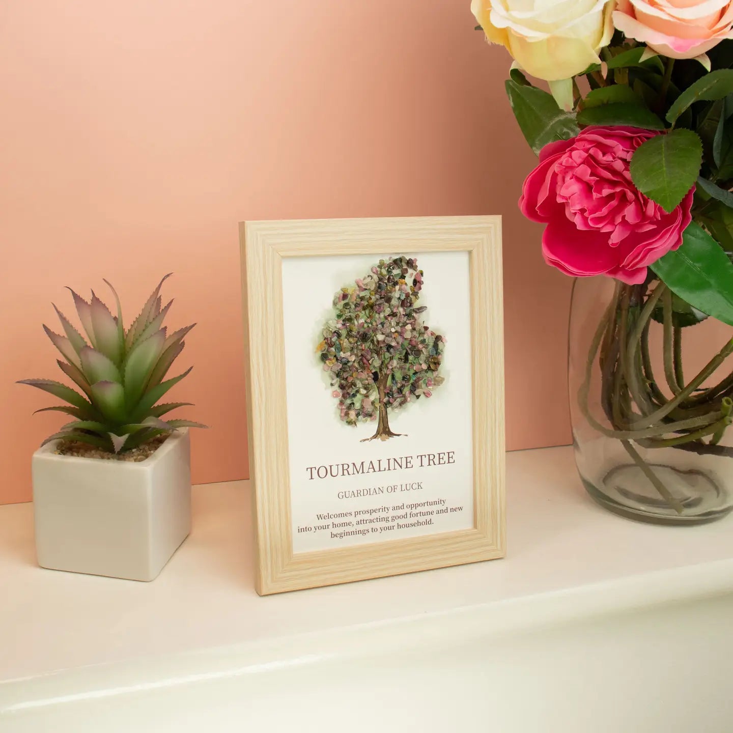 Framed artwork of a tree with text, placed on a surface with a plant and flowers against a peach wall.