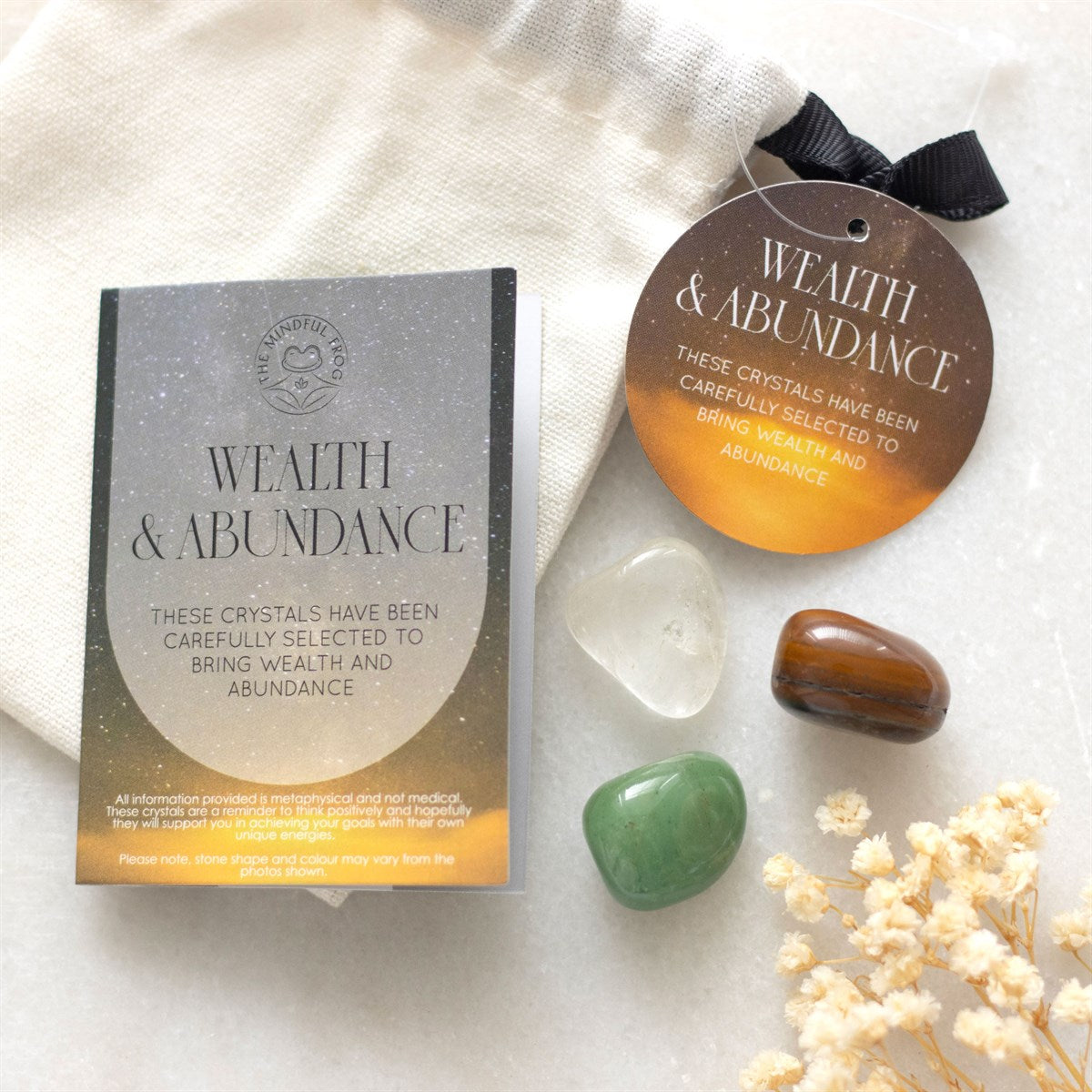 A set of healing crystals including green aventurine, clear quartz, and tiger's eye, presented in a drawstring bag with an informative insert card titled 'Wealth & Abundance'. The crystals are also accompanied by decorative dry plants and a black bow.