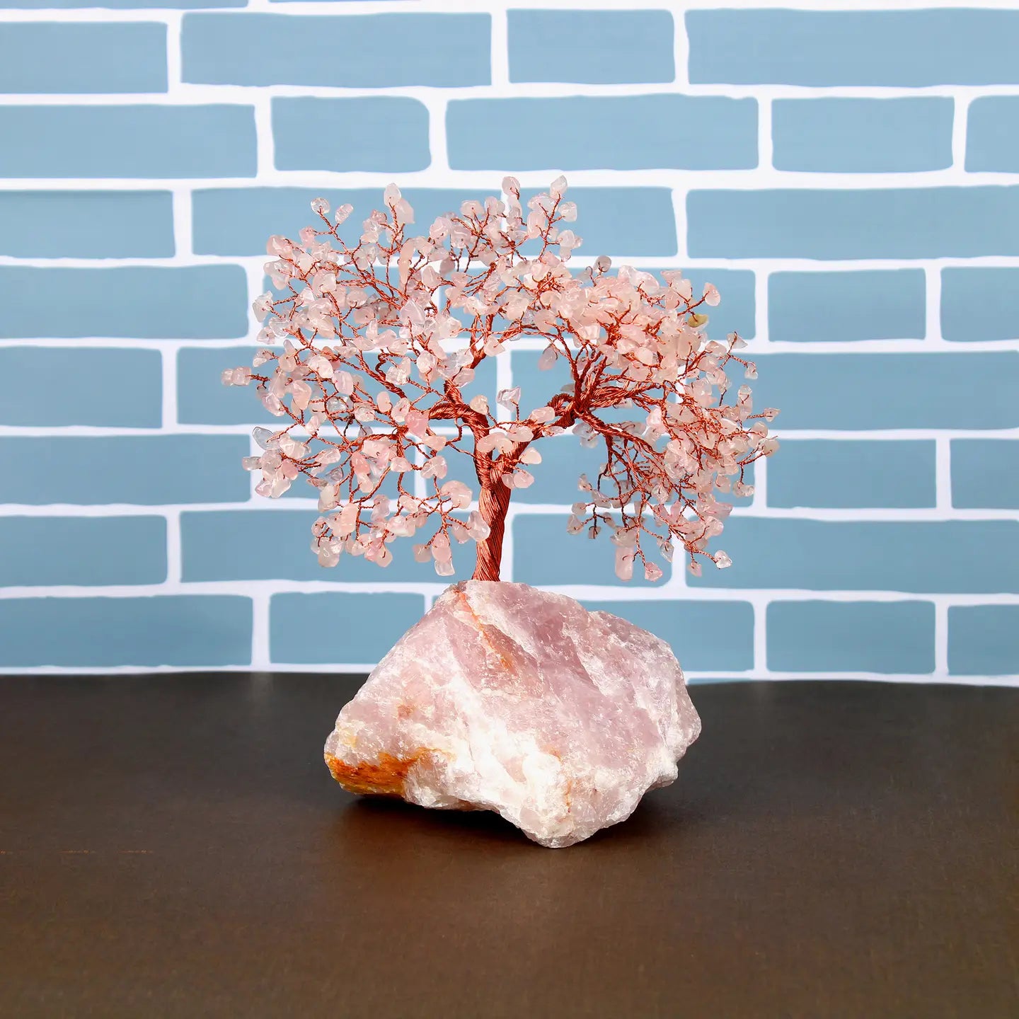 "Rose Quartz Crystal Tree with 500 beads in rose quartz base on brown countertop with blue brick background"